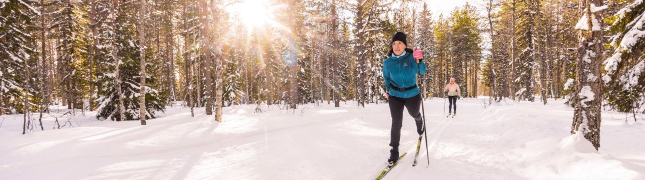 En kvinna med grön jacka åker längdskidor i en solig skog.