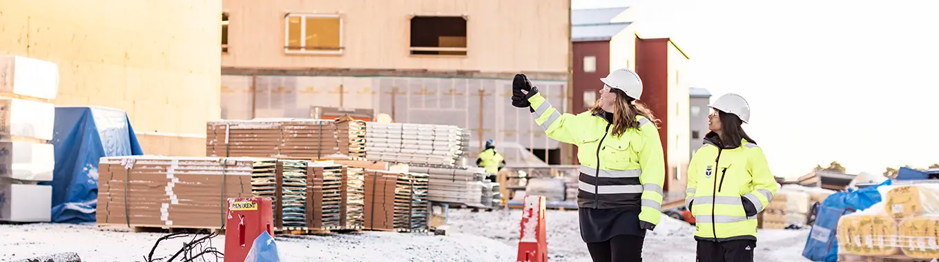 Två kvinnor med varseljackor och hjälmar går på ett bygge vintertid.