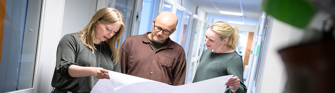 En man med glasögon och vinröd skjorta står i korridoren tillsammans med två kvinnliga kollegor och tittar på en stor ritning.