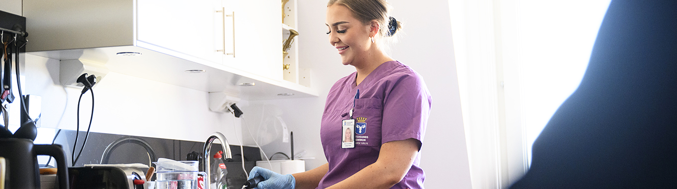 En ung kvinna som arbetar i hemtjänsten står i ett kök och diskar. Hon har mörkt hår och en lila arbetströja med kommunvapnet på.