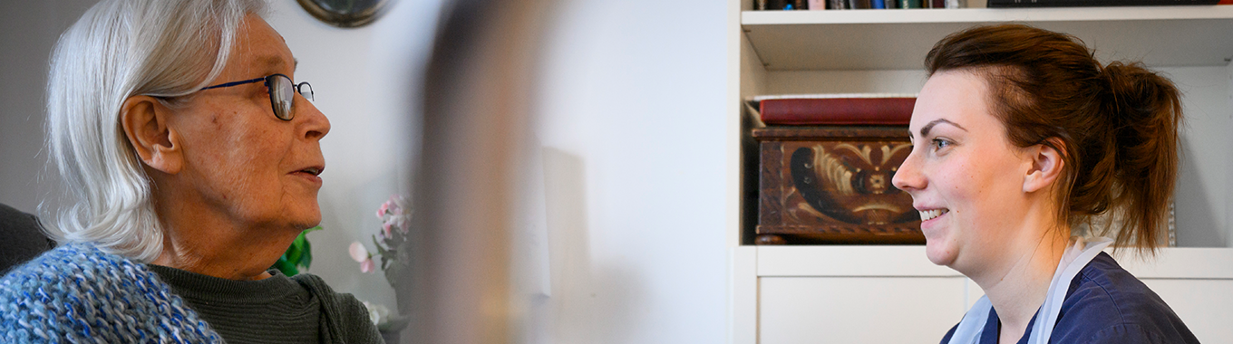 En yngre kvinna med mörkt hår och blå tröja sitter ner och hjälper en äldre kvinna  som har grått hår, page och glasögon. Den yngre kvinnan har ett förkläde på sig.